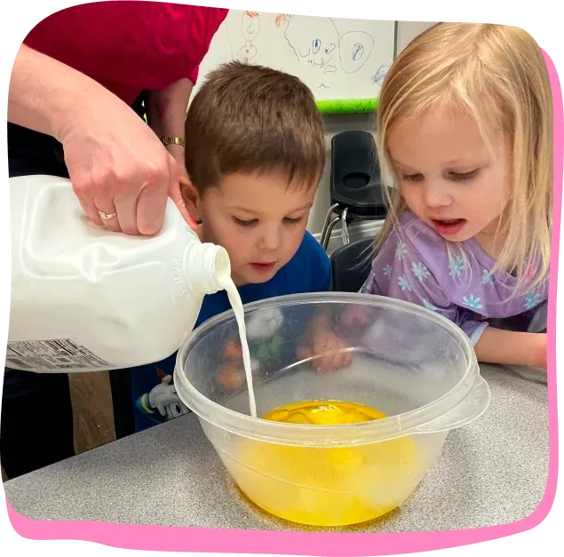 Two preschoolers watch a teacher pour milk into a mixing bowl at Songbird Academy