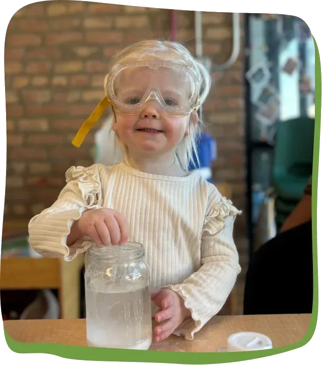 A preschooler wearing safety goggles holds a jar during a science experiment at Songbird Academy