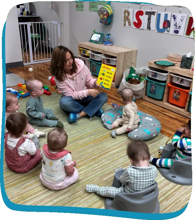 A teacher sits on the floor reading a picture book to babies in the Songbird Academy infant room